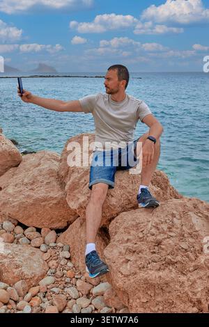 Ein junger Mann macht ein Selfie, während er auf großen Steinen am Meer sitzt Stockfoto