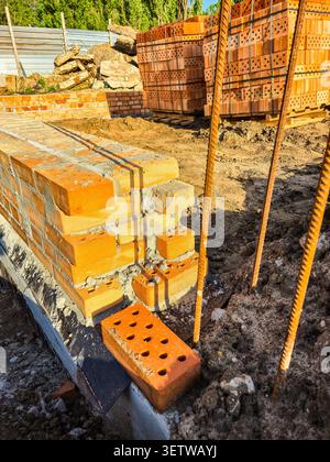 Auf einem Fundament mit Bewehrung und Ziegelsteinen in der Nähe einer Baustelle wird eine neue Ziegelmauer errichtet Stockfoto