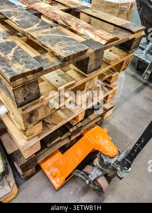 Eine Nahansicht zeigt einen Stapel verwitterter Holzpaletten mit einem orangefarbenen manuellen Palettenheber darunter, der für den Transport von Waren bereit ist Stockfoto