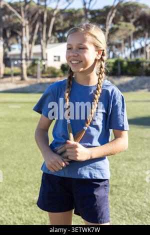 Ein junges Mädchen, das auf dem Grasfeld steht und Zöpfe hält, ein blaues Hemd und marineblaue Shorts in der Nähe des Tores trägt. Outdoor, sonnig, Jugend, sportlich, Lächeln, park, Bäume Stockfoto