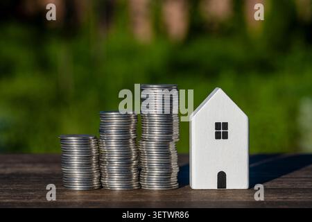 Weißes Miniaturhaus, neben wachsenden Silbermünzstapeln auf einem Holztisch, Nahaufnahme. Konzeptionelles Image für Immobilieninvestitionen, Hypothekenplanung, Stockfoto