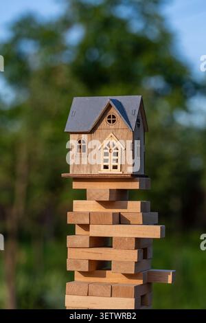 Holzhausmodell, das auf instablem Spielzeugblock-Turm balanciert. Das Konzept des Immobilienrisikos und der finanziellen Instabilität anhand eines kleinen Holzhausmodells Stockfoto