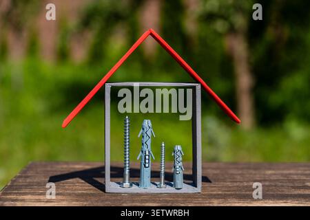 Kleiner symbolischer Hausrahmen gefüllt mit Bauschrauben und Kunststoff-Wanddübeln auf Holzoberfläche, Nahaufnahme. Konzeptbild für die Immobilienreparatur, Stockfoto