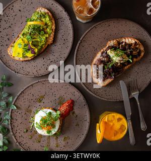 Ein flaches Gourmet-Frühstück mit Avocado-Toast, Pilz-Bruschetta, Maiskrapfen mit Spiegelei, Eislatte und Orange Stockfoto