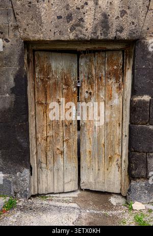 Alte Doppeltür aus Holz mit rostigen Metallscharnieren. Tür in Steinmauer, verwittert und veraltet. Gerissener Betonboden um die Basis Stockfoto