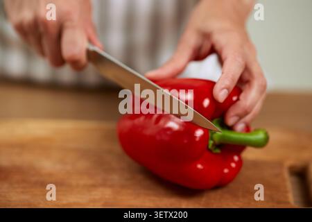Die Hände bereiten frische rote Paprika mit einem Kochmesser auf einem hölzernen Schneidebrett zu und machen gesunde Speisen Stockfoto