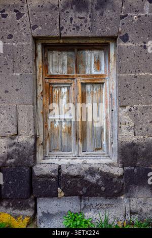 Altes Holzfenster in dunkler Steinmauer. Holz weist verwitterte, abblätternde Farbe und Rost auf. Steinblöcke um das Fenster sehen abgenutzt und moosig aus. Grüne Pflanzen g Stockfoto