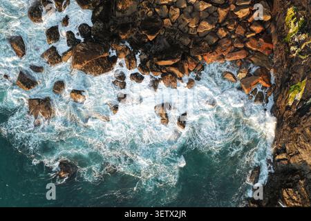 Von oben nach unten blickende weiße Wellen, die gegen dunkle felsige Klippen brechen und ein markantes Muster aus Schaum und Stein erzeugen. Stockfoto