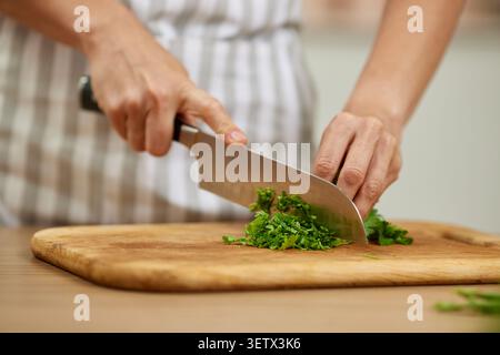 Hände hacken frische Petersilie oder Koriander mit einem scharfen Kochmesser auf einem hölzernen Schneidebrett, um Zutaten zum Kochen zuzubereiten Stockfoto