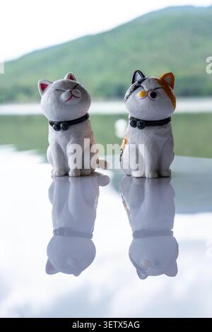 Chiang Mai, Thailand - 22. Juni 2026 : Weiße und Calico Katzenfiguren sitzen zusammen auf reflektierender Oberfläche mit lächelnden Ausdrücken und Bergrücken Stockfoto