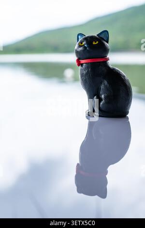 Chiang Mai, Thailand - 22. Juni 2026 : Schwarze Katzenfigur mit rotem Kragen auf reflektierender Oberfläche mit gelben Augen und Reservoir und Berg bac Stockfoto