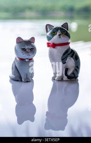 Chiang Mai, Thailand - 22. Juni 2026 : graue und tabbysche Katzenfiguren mit roten Kragen sitzen zusammen auf reflektierender Oberfläche mit unbeeinflusstem Ausdruck Stockfoto