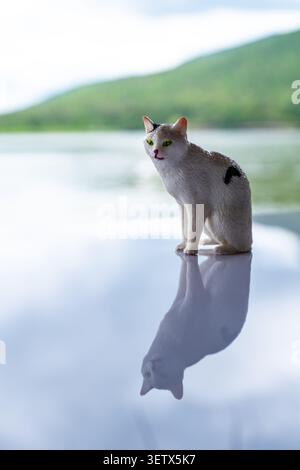 Chiang Mai, Thailand - 22. Juni 2026 : Weiße Katze Figur mit schwarzen Flecken auf reflektierender Oberfläche mit grünen Augen und Reservoir und Berg bac Stockfoto