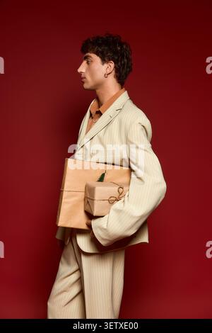 Ein junger Mann, elegant gekleidet, posiert selbstbewusst vor einem auffälligen roten Hintergrund und hält Geschenke. Stockfoto