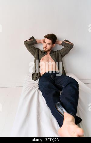 Ein junger Mann mit Bart entspannt sich auf einem Bett in einem Studio und zeigt seinen modischen Stil Stockfoto