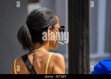 Seiten- und Rückansicht einer kubanischen Frau mit Sonnenbrille, einem Pferdeschwanz, einem kleinen Nackentattoo und einem Lutscher-Stab im Mund im Jose-Marti-Park in Ciego de Stockfoto