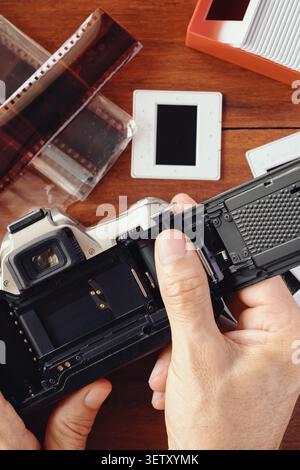 Nahaufnahme der Hände, die eine offene analoge Spiegelreflexkamera auf einem Holztisch mit Negativen, Diarahmen und Archivbox halten. Stockfoto