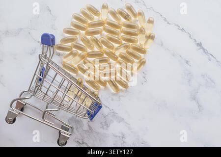 Hochwinkelansicht eines kleinen Einkaufswagens, der mit goldenen Fischöl-Gelkapseln auf einer weißen Marmoroberfläche umgekippt wurde und Medizin-pu konzeptualisiert Stockfoto