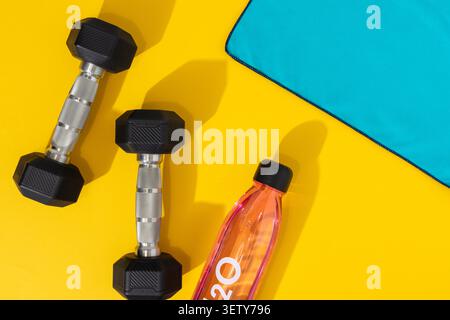 Flache Lage von zwei Sechskant-Kurzhanteln mit Wasserflasche und blauem Handtuch auf gelbem Hintergrund Stockfoto