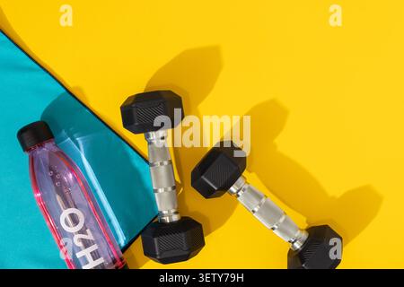 Flache Lage von zwei Sechskant-Kurzhanteln mit Wasserflasche und blauem Handtuch auf gelbem Hintergrund Stockfoto