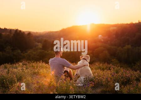 Mann sitzt zusammen mit seinem Hund auf grüner Graswiese während des wunderschönen goldenen Sonnenuntergangs. Freundschaft, Haustier-Begleitung, Outdoor-Relaxat Stockfoto