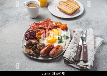 Komplettes englisches Frühstück auf einem Teller mit Spiegeleiern, knusprigem Speck, gegrillten Würstchen, Pilzen, Bohnen, Tomaten, Toast, Kaffee und Orangensaft. Stockfoto
