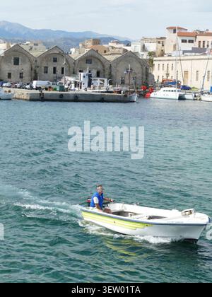 CHANIA, KRETA - 5. MAI 2025 - erfahrener Segler, der ein kleines Motorboot in der Nähe historischer venezianischer Arsenale im Hafen von Chania, Kreta, Griechenland, navigiert Stockfoto