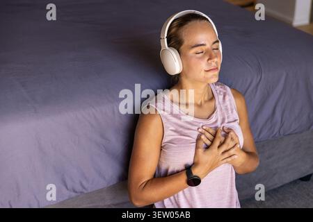 Frau Mitte 20, die neben dem Bett sitzt, mit grauer Federdecke, rosa Oberseite, Kopfhörern, Smartwatch, Kopierraum Stockfoto