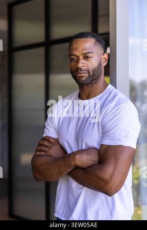 Reifer afroamerikanischer Mann, der sich auf einen Lichtrahmen im Eingang neben dunklem Glas stützt und weißes T-Stück trägt Stockfoto