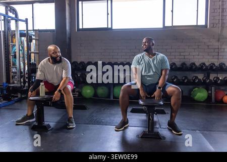 Zwei Afroamerikaner sitzen auf Bänken in Sportausrüstung und reden in der Nähe von Hanteln im Fitnessstudio Stockfoto