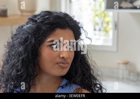 Indianerin sitzt in der Küche, trägt ärmelloses blaues Oberteil und blickt mit dem Glas zum Fenster Stockfoto