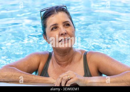 Eine ältere Frau, die sich ausruht und mit Kräuselung auf den Poolrand blickt, mit Badeanzug, Sonnenbrille, Ring Stockfoto