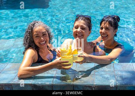 Verschiedene Freundinnen, die in dunklen Badeanzügen am Poolrand mit Zitrusgetränken und Sonnenbrille toasten Stockfoto