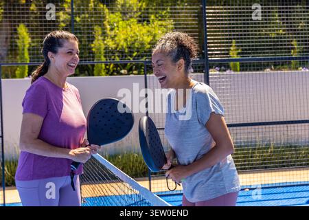 Verschiedene Freundinnen lachen und halten schwarze Paddel am Netz auf dem blauen Platz in sportlicher Ausrüstung Stockfoto
