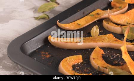 Scheiben gewürzter Butternusskürbis-Braten auf einem dunklen Blech mit Lorbeerblättern und Kräutern. Der Hochwinkelschuss fängt das goldene, karamellisierte Bild ein Stockfoto