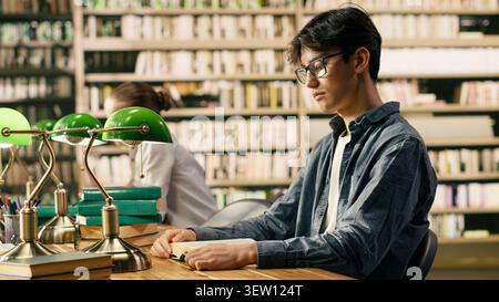 Junger asiatischer Mann mit Brille liest Buch am Studientisch der Bibliothek, zeigt fokussiertes akademisches Umfeld mit unabhängigem Studium. Lerninhalte für Schüler mit Kursmaterial und Lernmaterialien. Stockfoto