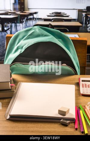 Türkisfarbener Rucksack auf einem Klassenstuhl hinter einem hölzernen Schreibtisch mit leerem Notizbuch, Büchern, rosa Taschenrechner, Buntstiften, Radiergummi und Spitzer. Stockfoto