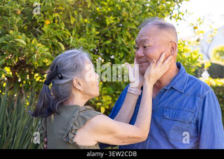 Asiatisches Seniorenpaar steht im sonnigen Garten, Frau schröpft das Gesicht des Mannes, Olivenbluse, Jeanshemd Stockfoto