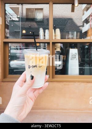 Eine Hand hält einen durchsichtigen Plastikbecher mit Eiskaffee vor einem japanischen Café, wobei das Schaufenster im Hintergrund sichtbar ist. Stockfoto