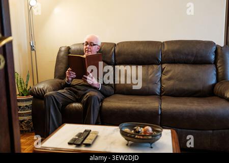 Ein älterer Mann, der sich im Sessel im Wohnzimmer entspannt, eine Brille trägt und ein Buch liest Stockfoto