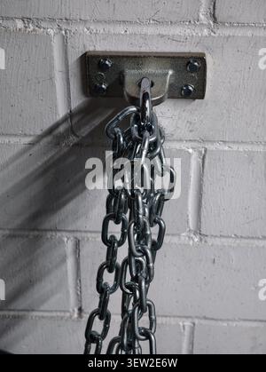 Nahaufnahme einer schweren Stahlkette, die an einer verschraubten Wandhalterung aus Metall aufgehängt ist, die an einer lackierten Ziegelwand montiert ist, Chromglieder reflektieren kühles Studiolicht, Prono Stockfoto