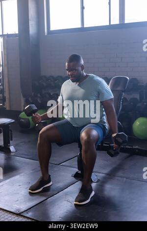 Afroamerikanischer Mann, der auf einer gepolsterten Bank sitzt und Hexenhanteln im Fitnessstudio an Ziegelwänden hält Stockfoto