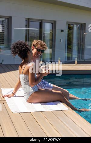 Diverse Paare sitzen am Pool auf Handtuch, Mann in Badehose, Frau in einteiligem Glas Stockfoto