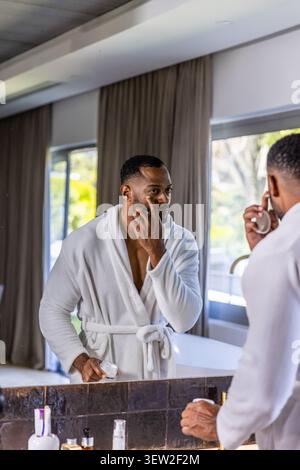 Reifer afroamerikanischer Mann, der am Kosmetikspiegel steht, in weißem Bademantel, der Creme aus dem Glas aufträgt Stockfoto
