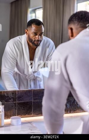 Reifer afroamerikanischer Mann mit weißem Bademantel, der sich am Waschbecken beugt und Spiegel studiert Stockfoto