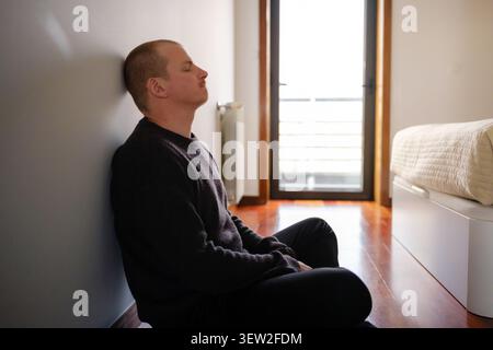 Mann sitzt auf dem Boden und lehnt sich an die Wand und findet Ruhe in einem hellen Raum Stockfoto