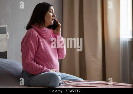 Schwangere Frau in rosa Pullover sitzt auf dem Bett, während sie drinnen am Telefon telefoniert Stockfoto