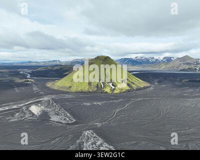 Aus der Vogelperspektive auf den lebhaften grünen Vulkan Maelifell, der aus den dunklen vulkanischen Sandebenen unter einem bewölkten Himmel in Vik, Myrdalshreppur, Island, aufsteigt. Stockfoto