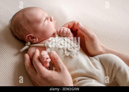 Erwachsene Hände halten das Neugeborene sanft auf einer weichen, gerippten Decke liegend. Das Baby blickt nach oben. Stockfoto