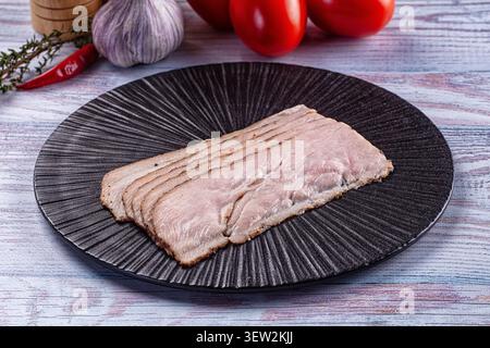 In Scheiben geschnittene Pastrami auf schwarzem geriffelten Teller mit leuchtenden roten Tomaten-Knoblauch-Chili im Hintergrund. Rustikale Holzoberfläche mit sanfter natürlicher Beleuchtung Stockfoto
